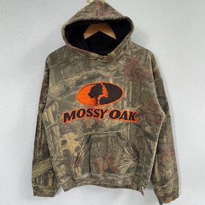 May include: A hooded sweatshirt with a camouflage pattern in shades of brown, green, and tan. The front features an orange oval with a tree silhouette and the text "MOSSY OAK" in orange. The hood has a black lining and a drawstring.