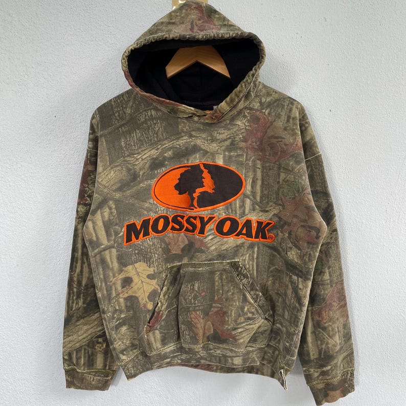 May include: A hooded sweatshirt with a camouflage pattern in shades of brown, green, and tan. The front features an orange oval with a tree silhouette and the text "MOSSY OAK" in orange. The hood has a black lining and a drawstring.