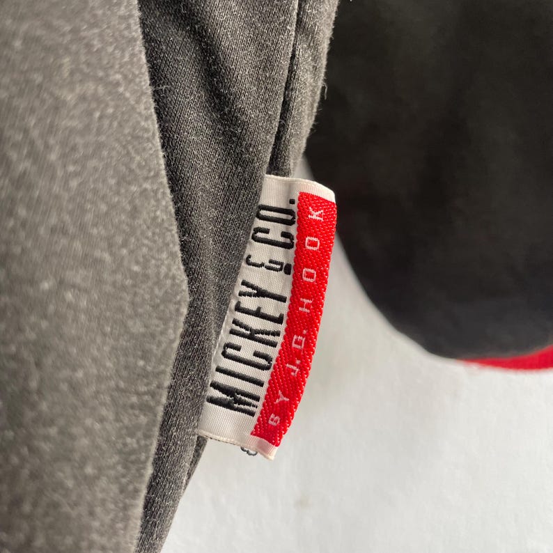 May include: Close-up of a gray t-shirt with a tag that reads "MICKEY & CO." in white text. The tag has a red border and the text "BY J.G. HOOK" in white. The shirt is partially visible, with a black section on the right.
