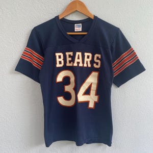 Puede incluir: Camiseta de fútbol vintage azul marino con la palabra "BEARS" en letras blancas y el número "34" en crema. Las mangas tienen rayas naranjas y blancas. La camiseta está colgada en una percha de madera.