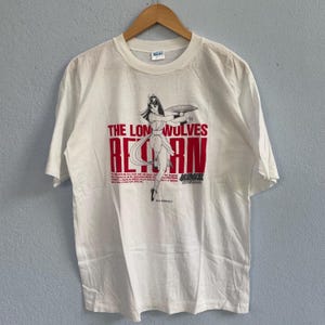 Puede incluir: Camiseta blanca con un gráfico de una mujer con un abanico y el texto rojo "THE LON WOLVES RETURN". La camiseta tiene mangas cortas y cuello redondo. El diseño incluye texto adicional y un logotipo.