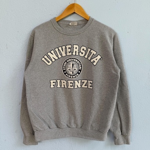 May include: A heather grey crewneck sweatshirt featuring the text UNIVERSITA' FIRENZE in white block lettering. A circular emblem sits in the centre, displaying a white fleur-de-lis symbol on a dark background with the text STUDI UNIVERSITARI.