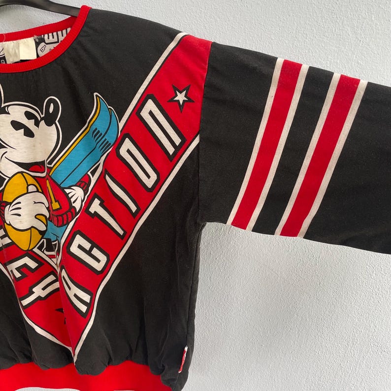 May include: Black long-sleeve sweatshirt featuring Mickey Mouse holding a football and a ski. The sweatshirt has red and white accents, with the word "ACTION" in white letters on a red banner. The sleeves have red and white stripes.