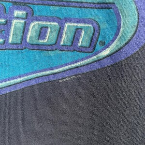 Op de afbeelding: Een donkerblauw T-shirt met een blauwe en witte afbeelding van het woord "Action" in een gestileerd lettertype. De afbeelding is omgeven door een blauwe ovale vorm. Het T-shirt heeft een kleine witte copyrightvermelding met de tekst "&copy;Xyla Arts, Inc."