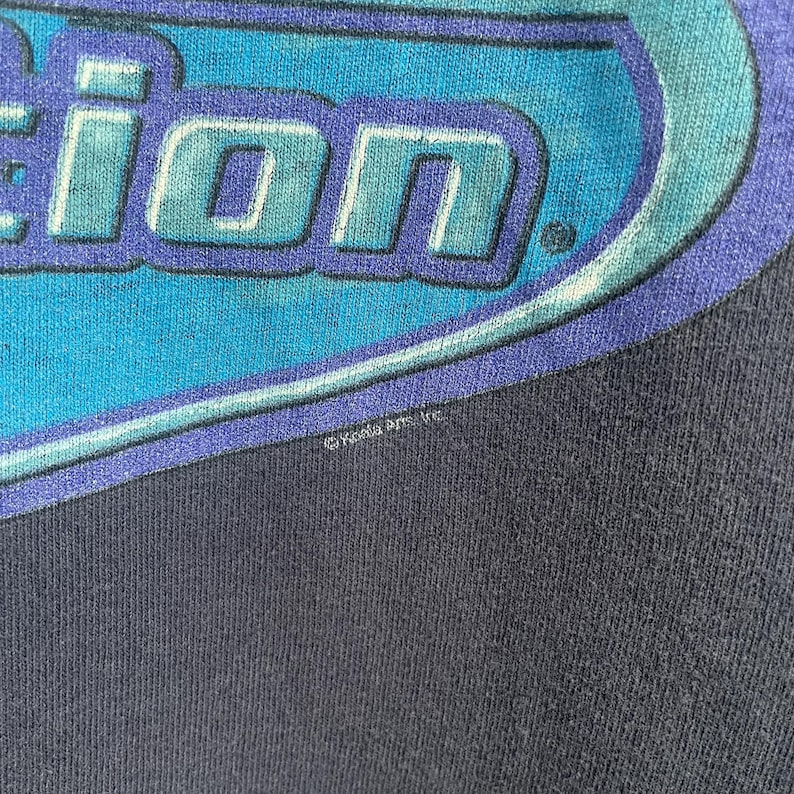 Op de afbeelding: Een donkerblauw T-shirt met een blauwe en witte afbeelding van het woord "Action" in een gestileerd lettertype. De afbeelding is omgeven door een blauwe ovale vorm. Het T-shirt heeft een kleine witte copyrightvermelding met de tekst "&copy;Xyla Arts, Inc."