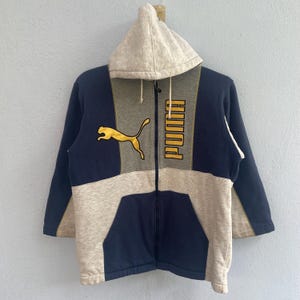Könnte beinhalten: Ein Kapuzen-Sweatshirt mit Reißverschluss in Marineblau, Grau und Beige. Das Sweatshirt hat das Puma-Logo in Gold auf der Brust und das Wort "PUMA" vertikal auf der Vorderseite. Die Jacke hat eine Vordertasche und eine Kapuze mit Kordelzug.