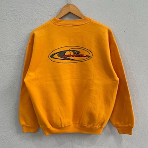 Puede incluir: Sudadera amarilla mostaza con el logo de O'Neill en la espalda. El logotipo tiene forma ovalada con la palabra "O'Neill" en rojo y negro. La sudadera está colgada en una percha de madera.