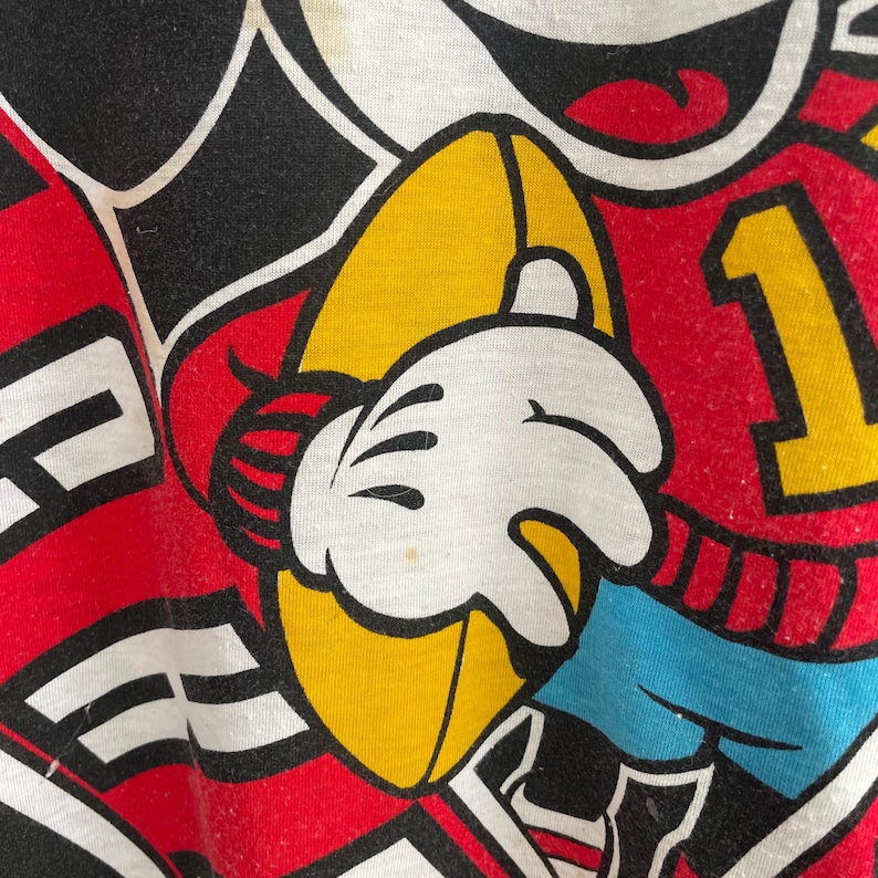 May include: Close-up of a white, yellow, red, and black graphic design. A white gloved hand is holding a yellow object. The number 1 is visible in yellow on a red background. The design is on a white background.