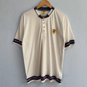 May include: Cream-colored polo shirt with a navy blue collar and sleeve trim featuring the word "Ferrari" in red. The shirt has a button-up placket and a yellow Ferrari logo on the chest.