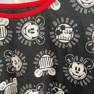 May include: Black t-shirt with a red neckline featuring a repeating pattern of Mickey Mouse and the words "Mickey Sports Club" in white. The design includes various Mickey Mouse images and text within circular and rectangular frames.
