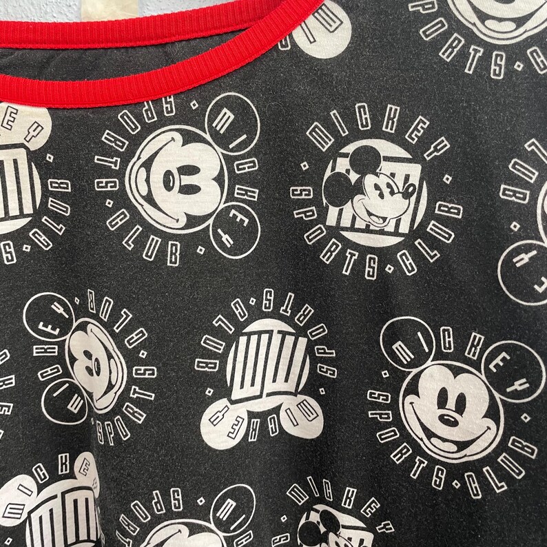 May include: Black t-shirt with a red neckline featuring a repeating pattern of Mickey Mouse and the words "Mickey Sports Club" in white. The design includes various Mickey Mouse images and text within circular and rectangular frames.