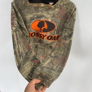 May include: A camouflage-patterned hoodie with the "Mossy Oak" logo in orange and black. The hoodie features a drawstring hood and a front pocket. The fabric has a mix of brown, green, and red tones.