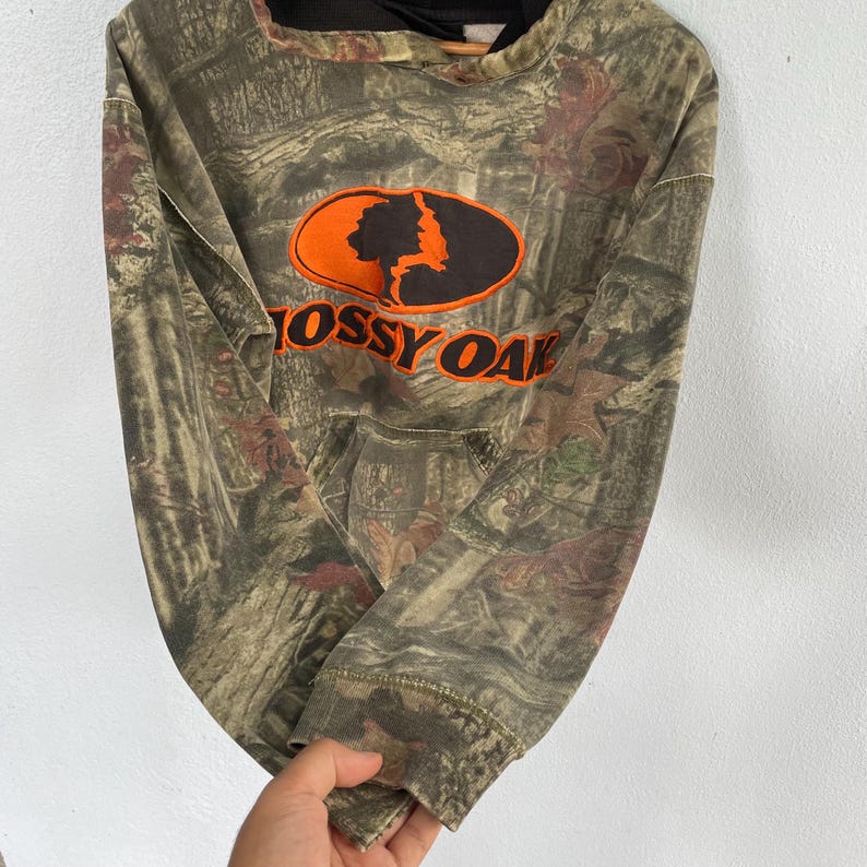 May include: A camouflage-patterned hoodie with the "Mossy Oak" logo in orange and black. The hoodie features a drawstring hood and a front pocket. The fabric has a mix of brown, green, and red tones.