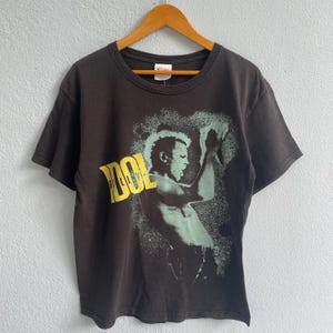 以下が含まれることがあります： 男性のグラフィックと黄色の「IDOL」の文字が入った黒いTシャツ。画像は、使い古されたヴィンテージの外観をしています。Tシャツは木製のハンガーに掛けられています。