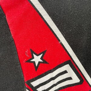 May include: Close-up of a black fabric with a red stripe and white accents. A white star is visible on the red stripe. The design is likely part of a larger garment or accessory.