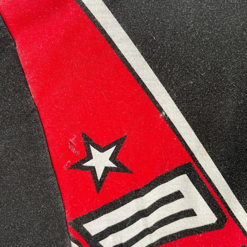 May include: Close-up of a black fabric with a red stripe and white accents. A white star is visible on the red stripe. The design is likely part of a larger garment or accessory.