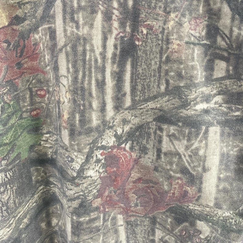 May include: Close-up of a fabric with a camouflage pattern. The design features a blend of brown, green, and gray tones, mimicking a forest environment. Reddish-brown leaf accents are scattered throughout the pattern.
