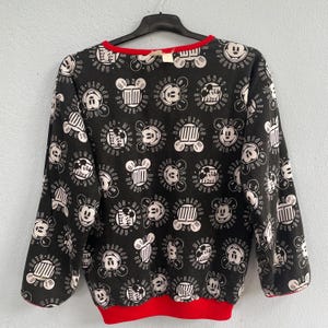 May include: Black sweatshirt with a repeating pattern of Mickey Mouse faces and other related graphics in white. The sweatshirt has a red neckline and cuffs. The garment is hanging on a black hanger.