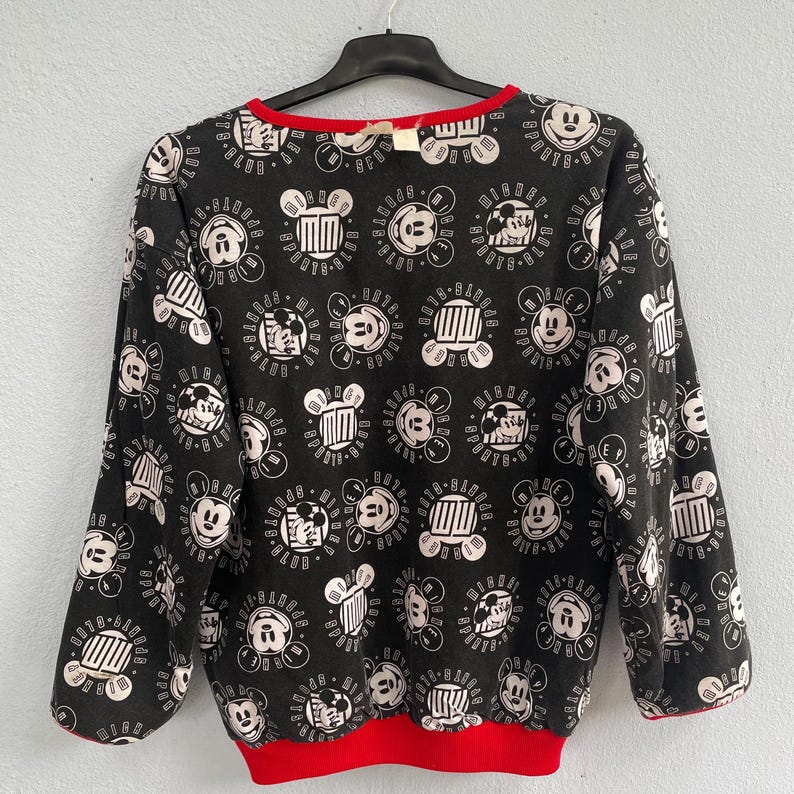 May include: Black sweatshirt with a repeating pattern of Mickey Mouse faces and other related graphics in white. The sweatshirt has a red neckline and cuffs. The garment is hanging on a black hanger.