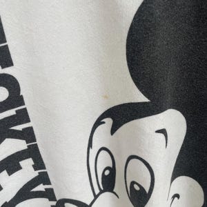 Puede incluir: Una prenda de tela blanca con un gran estampado gr&aacute;fico negro de la cabeza y las orejas de un personaje de dibujos animados. El texto MICKEY es parcialmente visible en letras may&uacute;sculas negras en el lado izquierdo de la tela, con una mancha.