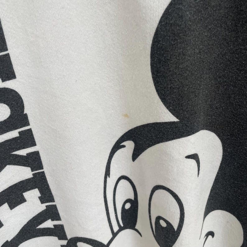 Puede incluir: Una prenda de tela blanca con un gran estampado gr&aacute;fico negro de la cabeza y las orejas de un personaje de dibujos animados. El texto MICKEY es parcialmente visible en letras may&uacute;sculas negras en el lado izquierdo de la tela, con una mancha.