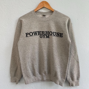 Può includere: Una felpa grigio melange con la scritta nera "POWERHOUSE GYM" stampata sul petto. La felpa è appesa a una gruccia di legno su uno sfondo di parete azzurra.