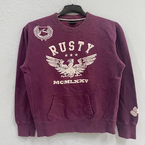 May include: A maroon sweatshirt with a front pocket and long sleeves. The front features the word "RUSTY" above a graphic of an eagle with outstretched wings. The text "MCMLXXV" is below the eagle. A logo patch is on the upper left chest.