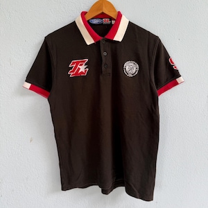 May include: A dark brown short-sleeve polo shirt featuring a red and white collar, a three-button placket, and red and white sleeve cuffs. The chest displays a red and white letter T logo and a circular crest patch with the text TL 1992.