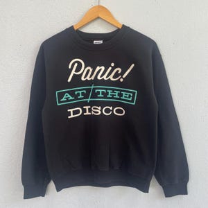 Könnte beinhalten: Schwarzes Sweatshirt mit dem Text "Panic! At The Disco" in Weiß und Türkis. Das Kleidungsstück hängt an einem Holzbügel vor einem hellgrauen Hintergrund. Der Text ist in einer stilisierten Schriftart gehalten.