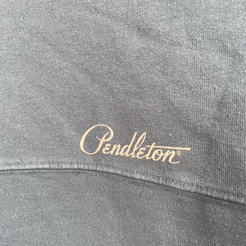 Może przedstawiać: Zbliżenie szarej bluzy z wyszytym słowem "Pendleton" w kursywie. Tekst jest w jasnobeżowym kolorze i jest wyśrodkowany na ubraniu. Tkanina ma teksturowany wygląd.