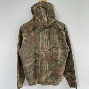 May include: A hooded sweatshirt with a camouflage pattern in shades of brown, green, and red. The hoodie is made of a soft material and features a drawstring hood and long sleeves. The back of the sweatshirt is visible.