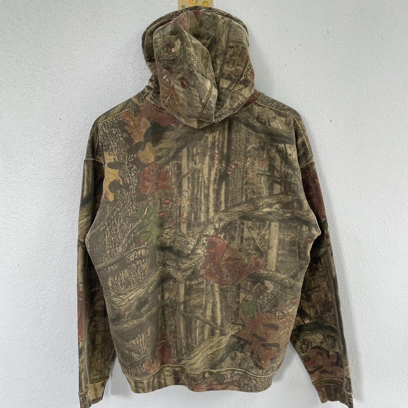 May include: A hooded sweatshirt with a camouflage pattern in shades of brown, green, and red. The hoodie is made of a soft material and features a drawstring hood and long sleeves. The back of the sweatshirt is visible.