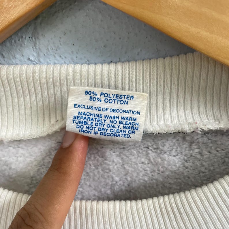 Puede incluir: Una etiqueta de prenda blanca con texto azul que dice 50% POLYESTER 50% COTTON, EXCLUSIVE OF DECORATION, MACHINE WASH WARM SEPARATELY. NO BLEACH. TUMBLE DRY ONLY, WARM. DO NOT DRY CLEAN OR IRON IF DECORATED. La etiqueta est&aacute; unida a un cuello blanco acanalado.