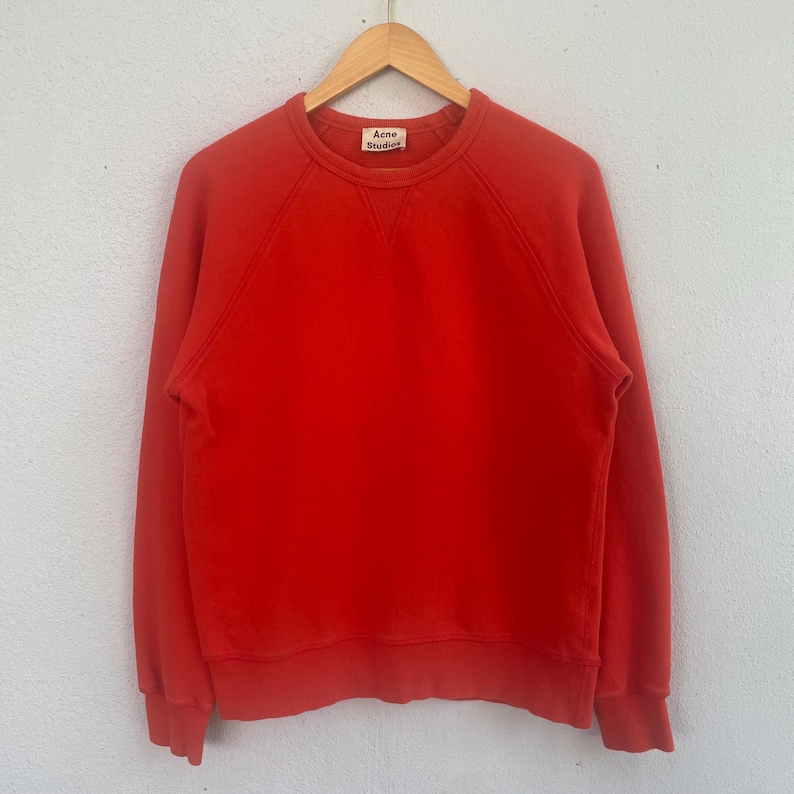 Op de afbeelding: Een effen rode sweatshirt met lange mouwen, hangend aan een houten hanger. De sweatshirt heeft een ronde hals en een klein label met de tekst "Acne Studios". Het kledingstuk is gemaakt van een zacht materiaal.
