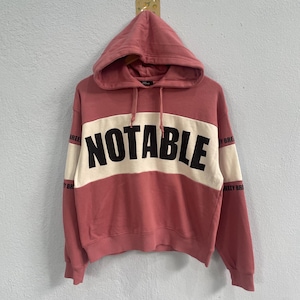 Peut inclure: Un sweat à capuche rose poudré avec une bande horizontale blanche sur la poitrine, portant le mot "NOTABLE" en grosses lettres noires. Les manches ont une bande crème et le mot "BREEZY" imprimé. Le sweat à capuche a un cordon de serrage et une capuche.
