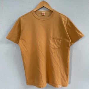 May include: Mustard-coloured short-sleeved t-shirt with a chest pocket. The shirt is hanging on a wooden hanger. The t-shirt has a crew neck and a small tag on the pocket and bottom hem.