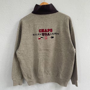 May include: A gray sweatshirt with a dark brown collar. The back of the sweatshirt features the embroidered text "CHAPS RALPH USA LAUREN" in red and white, along with nautical-themed graphics.