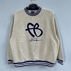 May include: A heather grey long-sleeve sweatshirt featuring a dark blue and white embroidered FB logo on the chest with the text Fubu below it. The garment has a ribbed crew neckline and cuffs with dark blue and white striped detailing.
