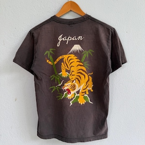 May include: A charcoal gray short-sleeve t-shirt featuring a graphic on the back. The design displays a roaring tiger, green bamboo stalks, and a white mountain peak above the word "japan" written in a white cursive script font.