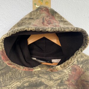 May include: A camouflage-patterned hoodie with a hood and a black interior. The hoodie is hanging on a wooden hanger. The camouflage design features shades of green, brown, and red.
