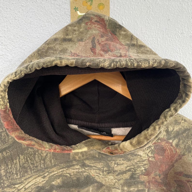 May include: A camouflage-patterned hoodie with a hood and a black interior. The hoodie is hanging on a wooden hanger. The camouflage design features shades of green, brown, and red.