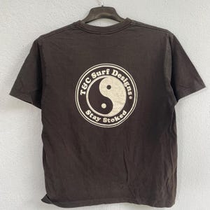 Puede incluir: Una camiseta marr&oacute;n con un gr&aacute;fico circular en la espalda. El gr&aacute;fico presenta un s&iacute;mbolo de yin-yang y el texto "T&C Surf Designs" y "Stay Stoked" en un dise&ntilde;o circular blanco. La camiseta est&aacute; colgada en una percha negra.