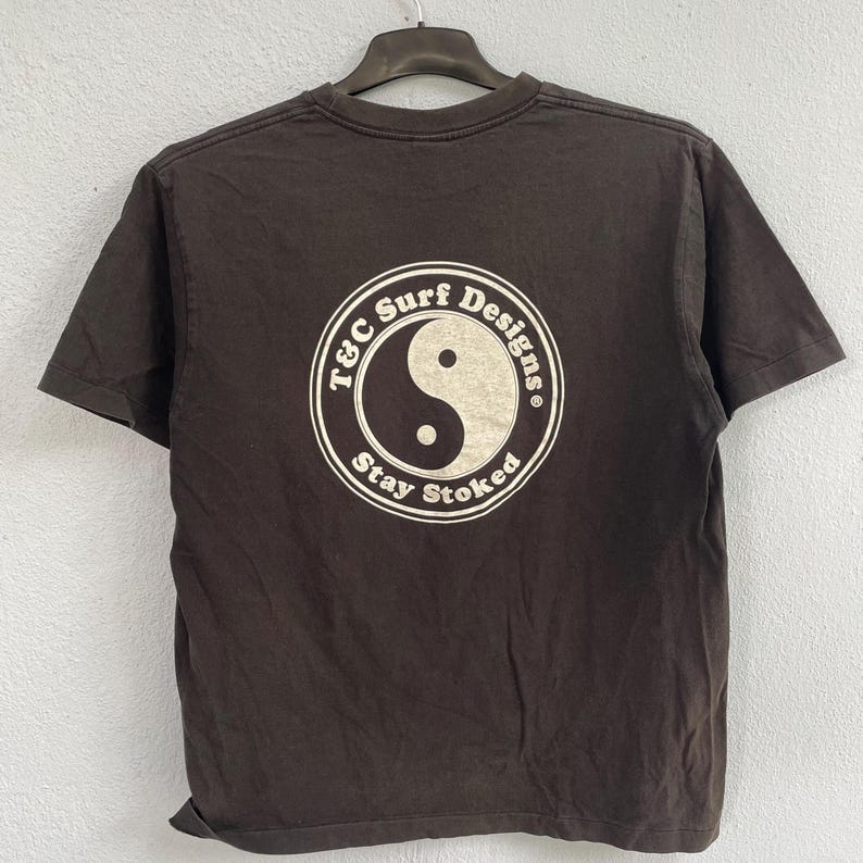 Puede incluir: Una camiseta marr&oacute;n con un gr&aacute;fico circular en la espalda. El gr&aacute;fico presenta un s&iacute;mbolo de yin-yang y el texto "T&C Surf Designs" y "Stay Stoked" en un dise&ntilde;o circular blanco. La camiseta est&aacute; colgada en una percha negra.
