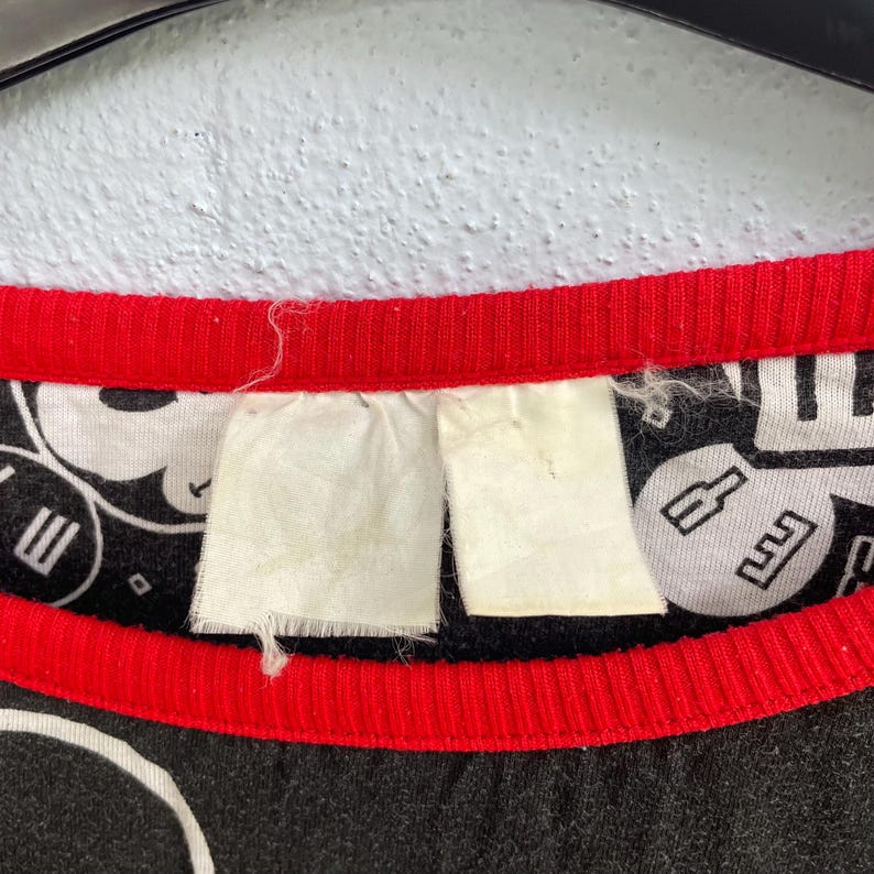 May include: Close-up of a black t-shirt collar with a red trim. The shirt features a white and black graphic print. Two white tags are attached to the collar. The shirt is hanging against a textured, off-white wall.