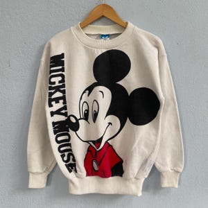 Puede incluir: Una sudadera de manga larga color crema con un gr&aacute;fico grande de Mickey Mouse en la parte delantera. El personaje aparece en negro, blanco y rojo, con el texto MICKEY MOUSE impreso verticalmente en letras negras negritas en el lado izquierdo.