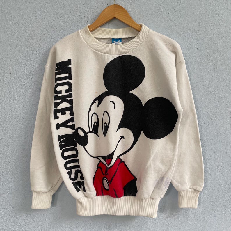 Puede incluir: Una sudadera de manga larga color crema con un gr&aacute;fico grande de Mickey Mouse en la parte delantera. El personaje aparece en negro, blanco y rojo, con el texto MICKEY MOUSE impreso verticalmente en letras negras negritas en el lado izquierdo.