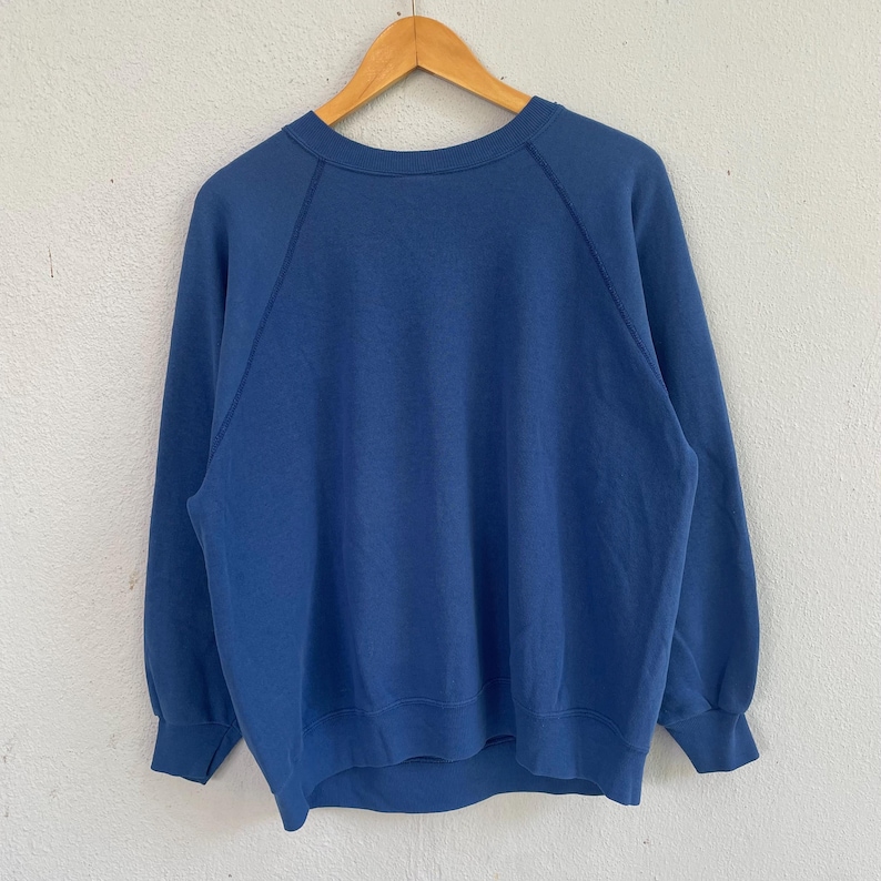 Peut inclure: Un sweat-shirt bleu uni &agrave; manches longues dot&eacute; d'une encolure ronde et de manches raglan. Le v&ecirc;tement est con&ccedil;u avec des poignets c&ocirc;tel&eacute;s aux poignets et un ourlet c&ocirc;tel&eacute; en bas, offrant une coupe structur&eacute;e. Le tissu a une texture lisse.