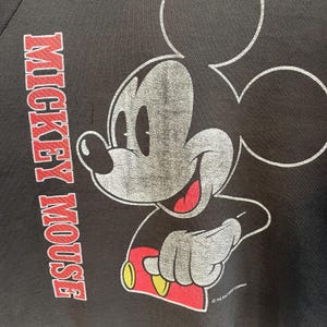 May include: Black sweatshirt with a distressed Mickey Mouse graphic. The design features Mickey's face and arm, with red, yellow, and grey details. The words "MICKEY MOUSE" are printed vertically in red on the left side of the garment.