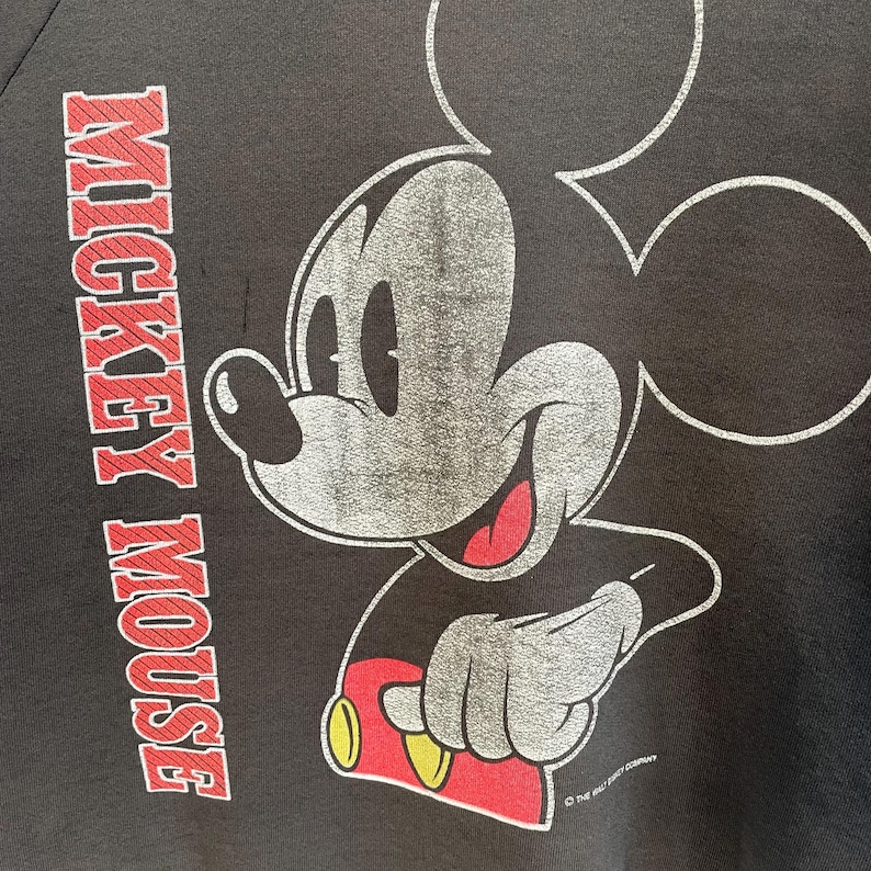 May include: Black sweatshirt with a distressed Mickey Mouse graphic. The design features Mickey's face and arm, with red, yellow, and grey details. The words "MICKEY MOUSE" are printed vertically in red on the left side of the garment.