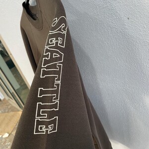 May include: A dark brown sweatshirt with the word "SEATTLE" printed vertically in white block letters on the left sleeve. The sweatshirt is hanging on a black hanger.